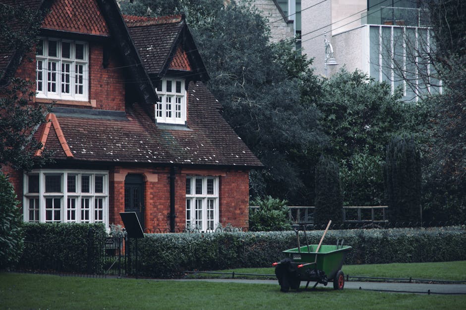 A person wearing a blue raincoat with a hood and black trousers, standing on a well-maintained lawn in a landscaped garden near Forty Hall Enfield, using a metal rake to clear fallen leaves and debris